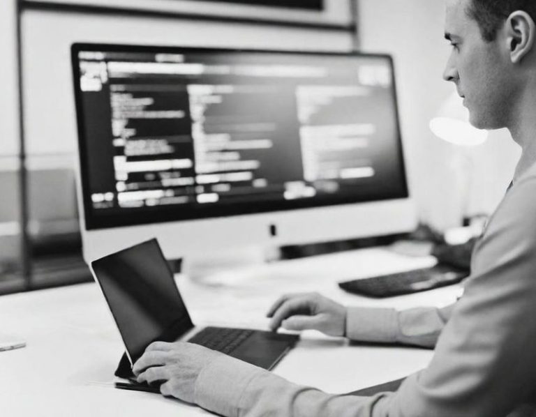 Mann mit Laptop vor einem großen Monitor, der Programmcode anzeigt.